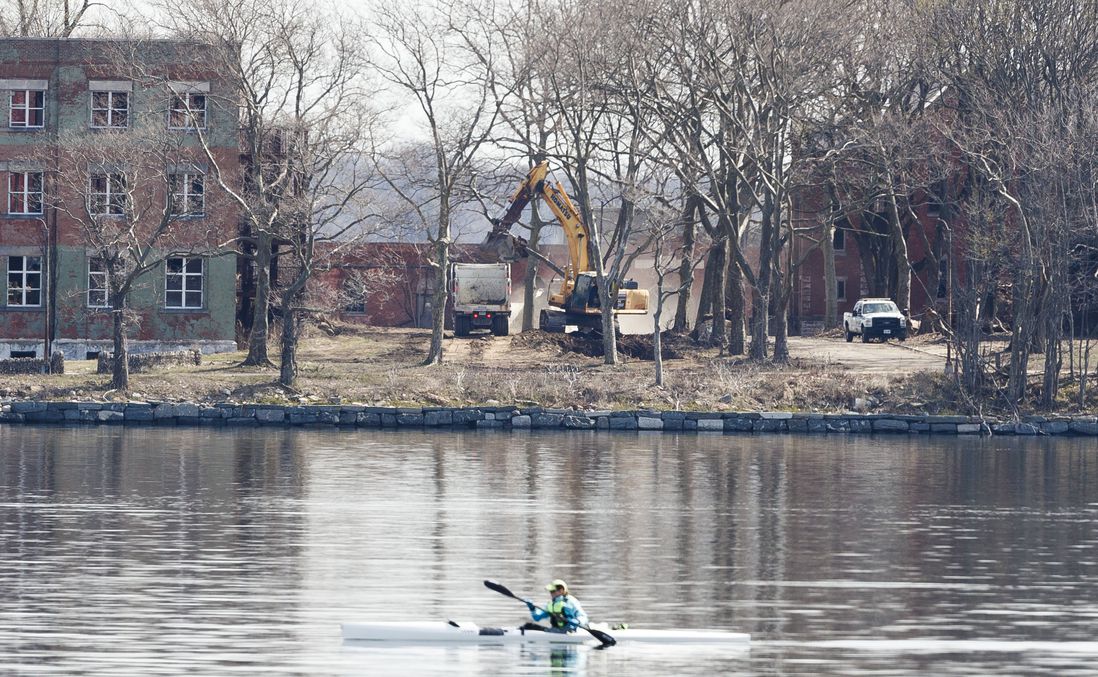 A backhoe digs a large hole and deposits dirt into dump trucks as a person in a kayak passes by in the foreground on Hart Island, located in the Long Island sound, off the coast of the Bronx, New York, USA, on 07 April 2020.